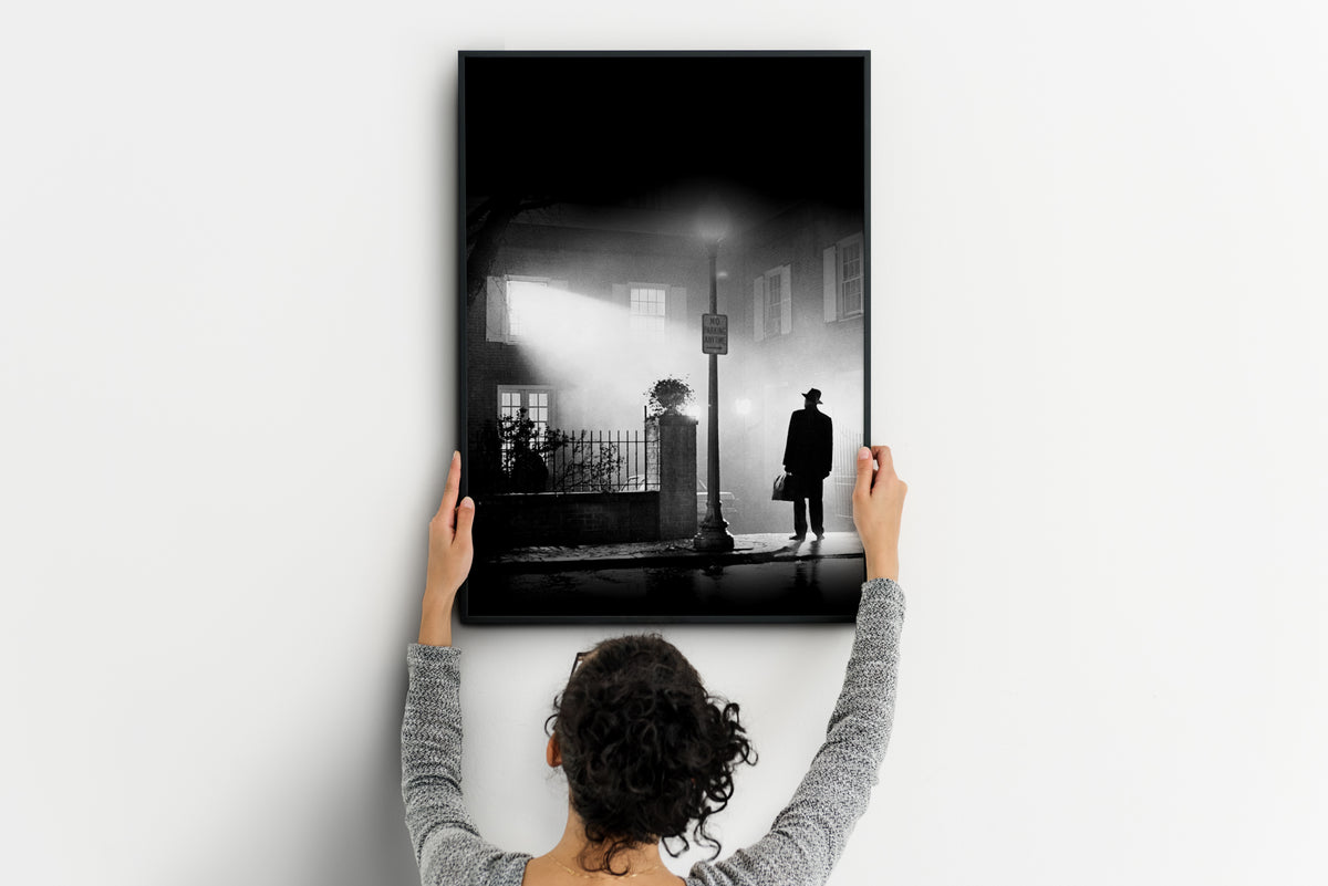 person holding framed black and white Exorcist movie poster with solitary figure in hat and coat on wet street at night