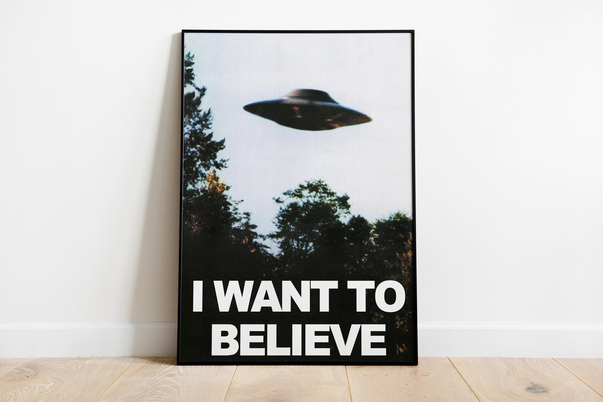 I Want to Believe UFO poster with a flying saucer above trees, shown in a moody living room interior.