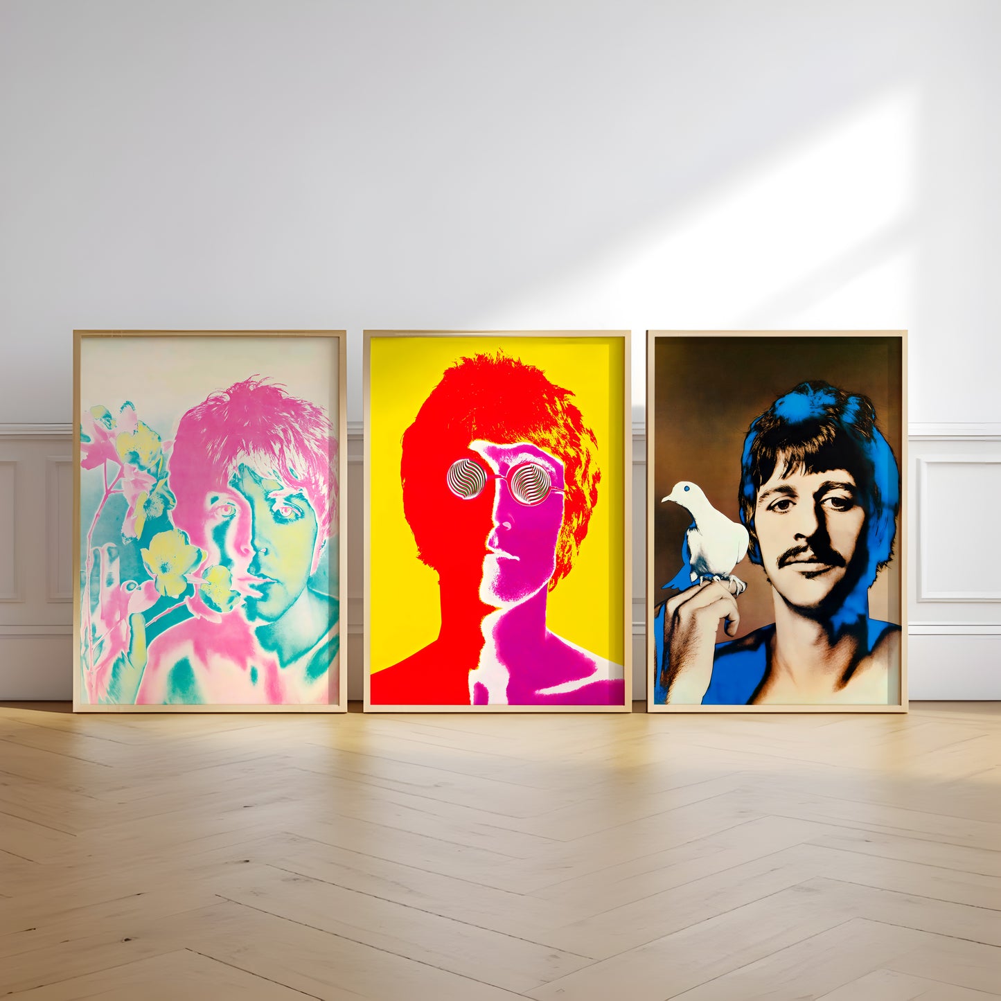 Three framed pop-art portraits of Beatles members John Lennon, Paul McCartney, and George Harrison displayed side-by-side on a light wood floor against a white wall.